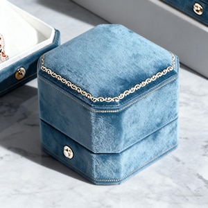 May include: A light blue velvet jewelry box with a gold trim and a gold clasp. The box is square with a hinged lid and sits on a marble surface. The box is empty.