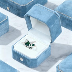 May include: A light blue velvet jewelry box with a silver-trimmed lid is open, revealing a ring with a green gemstone and silver accents. Several other closed boxes are visible in the background, suggesting a collection.