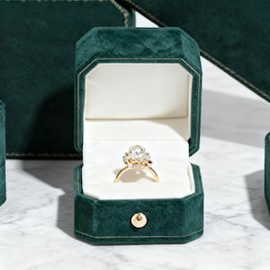 Puede incluir: Un anillo de compromiso de oro con una gema grande y transparente se muestra dentro de un joyero de terciopelo verde esmeralda abierto. El joyero tiene un interior blanco y un cierre de botón dorado. Otros joyeros son visibles en el fondo.