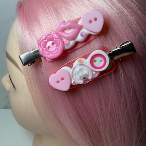 Op de afbeelding: Twee haarclips met een doorschijnende roze basis versierd met diverse ornamenten. De bovenste clip heeft een roze bloemknop, een roze lipvormige bedel en een lichtroze hartvormige knop. De onderste clip heeft een lichtroze hartvormige knop, een doorzichtige knop en een witte knop met een roze centrum.