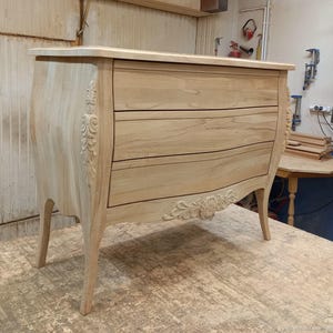 Puede incluir: Una cómoda de madera clara con tres cajones y tallas ornamentadas. La cómoda tiene lados y patas curvados y está sin terminar. La parte superior de la cómoda es plana.