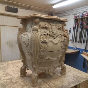 Puede incluir: Una taza de cerámica blanca con asa negra y el texto "But First, Coffee" en negrita negra. La taza se encuentra sobre un fondo borroso, sugiriendo un ambiente acogedor.