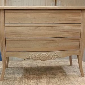 Puede incluir: Una cómoda de madera clara con tres cajones y tallas ornamentales. La cómoda tiene patas curvas y detalles decorativos en los laterales e inferior. La pieza está sin terminar y parece estar en un taller.
