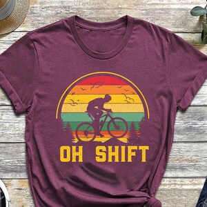 May include: Burgundy t-shirt with a bicycle rider graphic inside a sunset design. The text "OH SHIFT" is below the graphic. The shirt is displayed with black sneakers, a straw hat, glasses, and blue jeans.