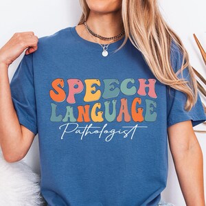 May include: Indigo blue t-shirt with the words "SPEECH LANGUAGE Pathologist" in colorful, retro-style lettering. The shirt has a crew neck and short sleeves. The text is in orange, pink, green, and yellow.