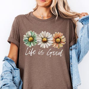 Puede incluir: Camiseta marr&oacute;n jaspeado con tres flores de margarita en verde, blanco y naranja con centros amarillos. Las palabras "Life is Good" est&aacute;n escritas en cursiva debajo de las flores. La camiseta tiene mangas enrolladas.
