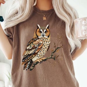 May include: Brown t-shirt featuring a detailed illustration of an owl perched on a branch. The owl has orange and brown feathers with intricate patterns. The shirt is a solid brown color.