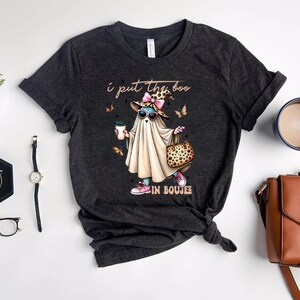 May include: A dark heather gray t-shirt featuring a graphic of a ghost wearing a leopard print hat with a pink bow, sunglasses, and pink sneakers. The ghost holds a coffee cup and a leopard print handbag. The text reads, I put the boo IN BOUJEE.