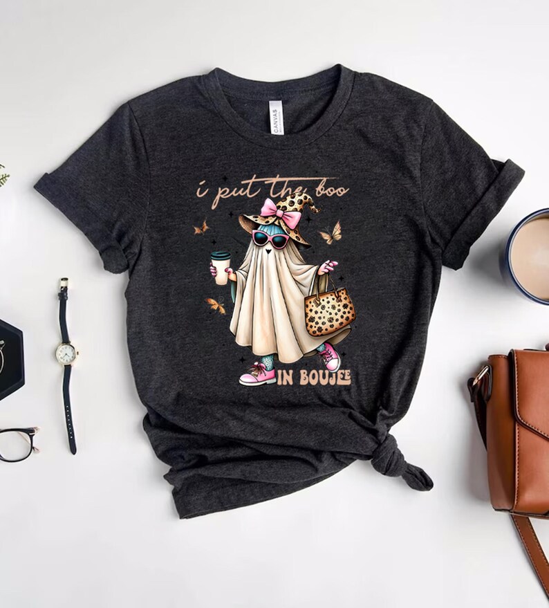 May include: A dark heather gray t-shirt featuring a graphic of a ghost wearing a leopard print hat with a pink bow, sunglasses, and pink sneakers. The ghost holds a coffee cup and a leopard print handbag. The text reads, I put the boo IN BOUJEE.