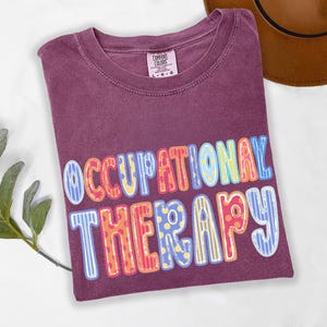 May include: A plum-colored short-sleeve t-shirt with a crew neckline and a Comfort Colors brand label. The front features the text OCCUPATIONAL THERAPY in large, multi-colored, patterned block letters with stripes and polka dots in blue, yellow, and red.