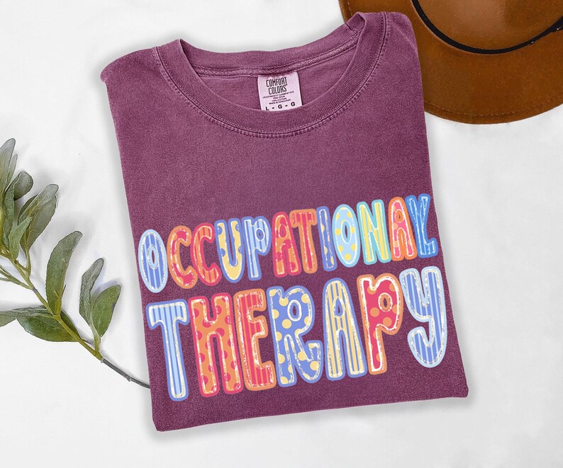 May include: A plum-colored short-sleeve t-shirt with a crew neckline and a Comfort Colors brand label. The front features the text OCCUPATIONAL THERAPY in large, multi-colored, patterned block letters with stripes and polka dots in blue, yellow, and red.
