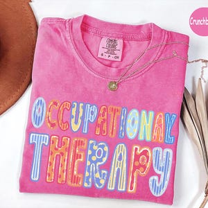 May include: A bright pink short-sleeved t-shirt with a crew neckline and a Comfort Colors label. The front features the text OCCUPATIONAL THERAPY in large, multi-colored, patterned block letters. A delicate gold-toned necklace rests on the shirt.