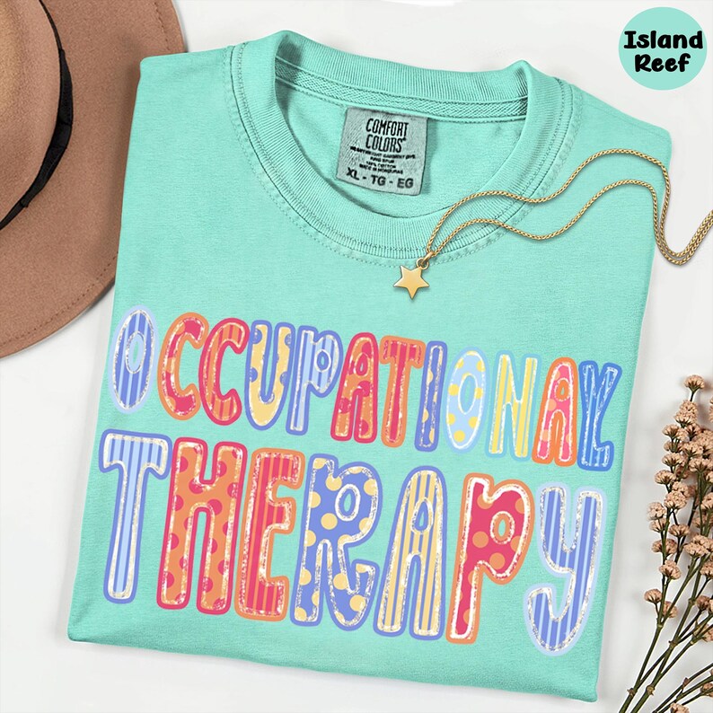 May include: A mint green t-shirt with a Comfort Colors label, featuring the text OCCUPATIONAL THERAPY in large, multi-colored, patterned block letters. The shirt is accented by a gold-toned necklace with a small star pendant.