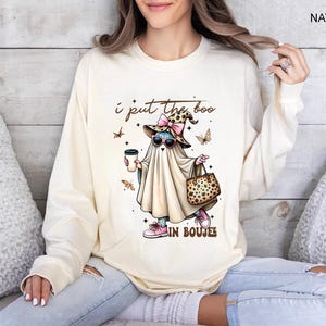 May include: A natural-colored long-sleeve sweatshirt featuring a graphic of a ghost wearing a leopard print witch hat with a pink bow, pink sunglasses, and pink sneakers. The ghost holds a coffee cup and a leopard print bag with text I put the boo in boujee.