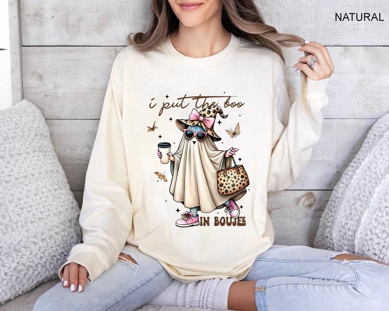 May include: A natural-colored long-sleeve sweatshirt featuring a graphic of a ghost wearing a leopard print witch hat with a pink bow, pink sunglasses, and pink sneakers. The ghost holds a coffee cup and a leopard print bag with text I put the boo in boujee.