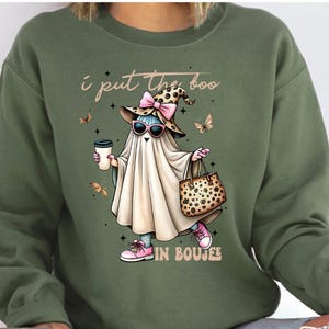 May include: An olive green crewneck sweatshirt featuring a graphic of a ghost wearing a leopard print witch hat with a pink bow, pink sunglasses, and pink sneakers. The ghost holds a coffee cup and a leopard print handbag, with text reading I put the boo IN BOUJEE.
