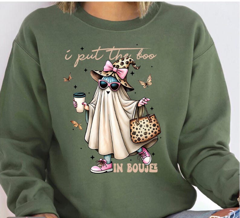 May include: An olive green crewneck sweatshirt featuring a graphic of a ghost wearing a leopard print witch hat with a pink bow, pink sunglasses, and pink sneakers. The ghost holds a coffee cup and a leopard print handbag, with text reading I put the boo IN BOUJEE.