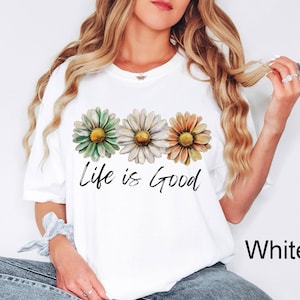 Puede incluir: Camiseta blanca con tres margaritas en tonos verdes, blancos y naranjas, con centros amarillos. La frase "Life is Good" est&aacute; escrita en negro debajo. La camiseta es de estilo casual.