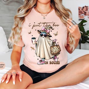 May include: A heather peach t-shirt featuring a graphic of a ghost wearing a leopard print hat with a pink bow, sunglasses, and pink sneakers. The ghost holds a coffee cup and a leopard print handbag. The text reads, I put the boo in BOUJEE.