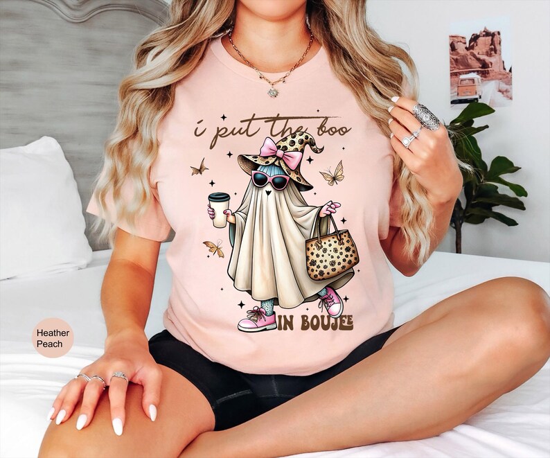 May include: A heather peach t-shirt featuring a graphic of a ghost wearing a leopard print hat with a pink bow, sunglasses, and pink sneakers. The ghost holds a coffee cup and a leopard print handbag. The text reads, I put the boo in BOUJEE.