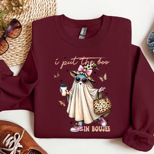 May include: A maroon long-sleeve sweatshirt featuring a graphic of a ghost wearing a leopard print witch hat, pink sunglasses, and pink sneakers, holding a coffee cup and a leopard print handbag, with the text I put the boo in boujee.