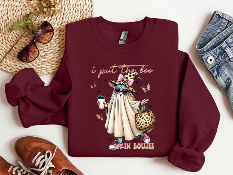May include: A maroon long-sleeve sweatshirt featuring a graphic of a ghost wearing a leopard print witch hat, pink sunglasses, and pink sneakers, holding a coffee cup and a leopard print handbag, with the text I put the boo in boujee.