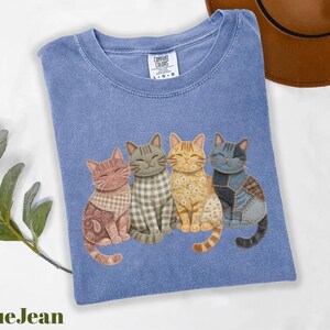 K&ouml;nnte beinhalten: Blaues T-Shirt mit vier Cartoon-Katzen mit einzigartigen Mustern und Farben. Die Katzen sitzen nebeneinander, jede mit einem unverwechselbaren Design, darunter Paisley, Karomuster und Patchwork. Das Shirt hat einen Rundhalsausschnitt und ein kleines Etikett oben.