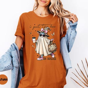 May include: A burnt orange t-shirt featuring a graphic of a ghost wearing a leopard print witch hat, pink sunglasses, and pink sneakers. The ghost holds a coffee cup and a leopard print handbag. The text reads I put the boo in boujee.