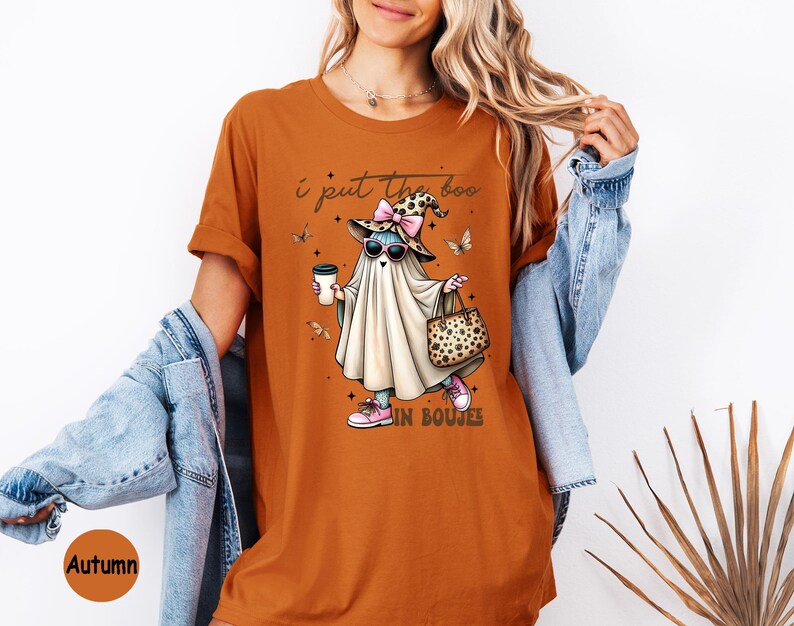 May include: A burnt orange t-shirt featuring a graphic of a ghost wearing a leopard print witch hat, pink sunglasses, and pink sneakers. The ghost holds a coffee cup and a leopard print handbag. The text reads I put the boo in boujee.