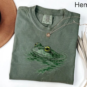 May include: A hemp-colored short-sleeve t-shirt featuring a graphic print of a frog head emerging from water. The frog has prominent yellow eyes and green skin. The shirt has a crew neckline and a Comfort Colors brand label on the inside collar.