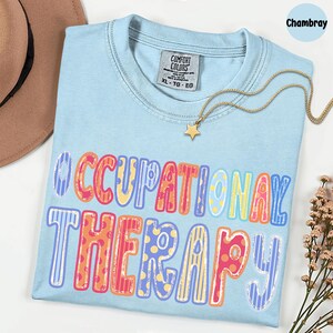 May include: A light blue crew neck t-shirt featuring the text OCCUPATIONAL THERAPY in a multi-colored, patterned font. The shirt has a Comfort Colors label in the collar, size XL, and is displayed with a gold star pendant necklace.