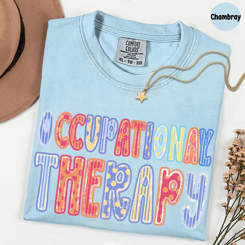 May include: A light blue crew neck t-shirt featuring the text OCCUPATIONAL THERAPY in a multi-colored, patterned font. The shirt has a Comfort Colors label in the collar, size XL, and is displayed with a gold star pendant necklace.