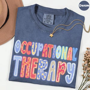 May include: A denim blue t-shirt featuring the text OCCUPATIONAL THERAPY in large, multi-colored letters with various patterns including stripes and polka dots. The shirt has a Comfort Colors label at the neck and is paired with a gold star necklace.