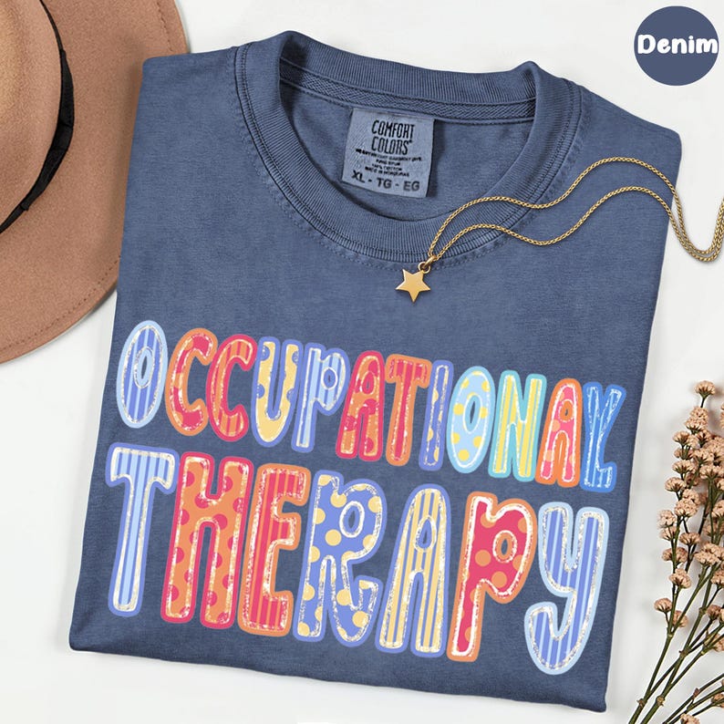May include: A denim blue t-shirt featuring the text OCCUPATIONAL THERAPY in large, multi-colored letters with various patterns including stripes and polka dots. The shirt has a Comfort Colors label at the neck and is paired with a gold star necklace.