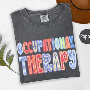 May include: A charcoal gray t-shirt with the text OCCUPATIONAL THERAPY printed in a multi-colored, patterned font featuring stripes and polka dots in shades of blue, yellow, orange, and pink. The shirt has a Comfort Colors brand neck label.