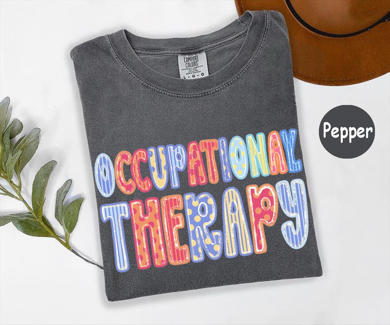 May include: A charcoal gray t-shirt with the text OCCUPATIONAL THERAPY printed in a multi-colored, patterned font featuring stripes and polka dots in shades of blue, yellow, orange, and pink. The shirt has a Comfort Colors brand neck label.