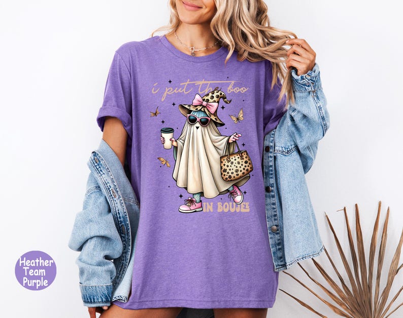 May include: A heather purple t-shirt featuring a graphic of a ghost wearing a leopard print witch hat, pink sunglasses, and pink sneakers. The ghost holds a coffee cup and a leopard print handbag. The text reads, I put the boo in boujee.