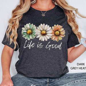 Puede incluir: Camiseta gris jaspeado oscuro con tres flores de margarita en verde, blanco y naranja con centros amarillos. Las palabras "Life is Good" est&aacute;n impresas en blanco debajo. Una prenda informal para el d&iacute;a a d&iacute;a.