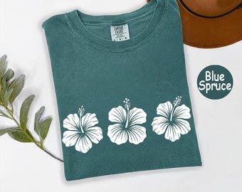 May include: A blue spruce t-shirt with three white hibiscus flower designs. The shirt is a solid color with a crew neck and short sleeves. The text "Blue Spruce" is in a circle on the right side of the shirt.