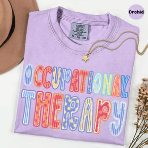 May include: A light purple short-sleeve t-shirt with a crew neckline and a Comfort Colors label. The front features the text OCCUPATIONAL THERAPY in large, multi-colored letters with various patterns including polka dots and vertical stripes.