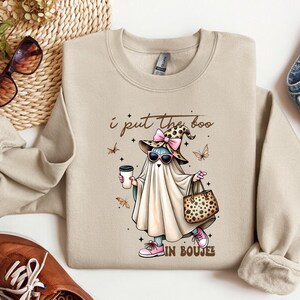 May include: A beige crewneck sweatshirt featuring a graphic of a ghost wearing a leopard print witch hat with a pink bow, sunglasses, and pink sneakers. The ghost holds a coffee cup and a leopard print handbag. The text reads I put the boo in boujee.