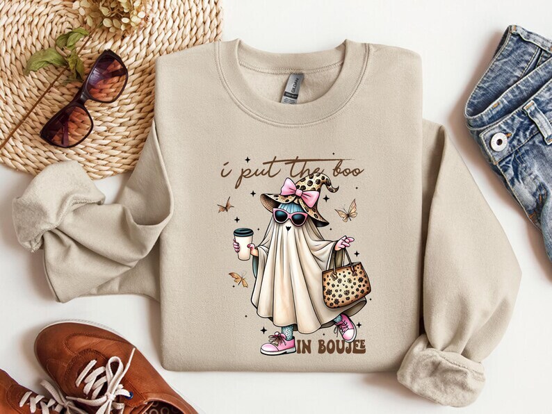 May include: A beige crewneck sweatshirt featuring a graphic of a ghost wearing a leopard print witch hat with a pink bow, sunglasses, and pink sneakers. The ghost holds a coffee cup and a leopard print handbag. The text reads I put the boo in boujee.