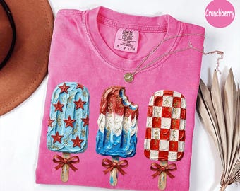Może przedstawiać: Różowa koszulka Comfort Colors z trzema wzorami lodów na patyku. Jeden jest niebieski z czerwonymi gwiazdami, drugi czerwono-biało-niebieski z kęsem, a ostatni w czerwono-białą kratę. Każdy lód na patyku ma drewniany patyczek i czerwoną kokardę.