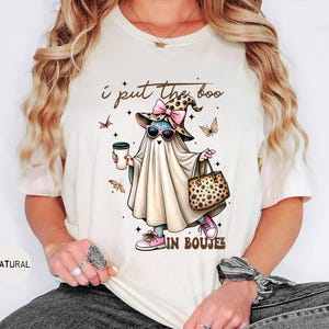 May include: A natural-colored t-shirt featuring a graphic of a ghost wearing a leopard print witch hat with a pink bow, pink sunglasses, and pink sneakers. The ghost holds a coffee cup and a leopard print handbag. Text reads: I put the boo in boujee.
