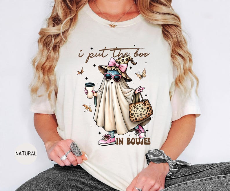 May include: A natural-colored t-shirt featuring a graphic of a ghost wearing a leopard print witch hat with a pink bow, pink sunglasses, and pink sneakers. The ghost holds a coffee cup and a leopard print handbag. Text reads: I put the boo in boujee.