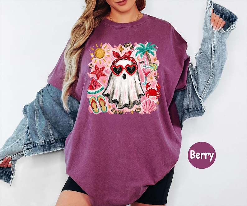 May include: A berry-colored t-shirt featuring a graphic print of a ghost wearing a red bandana and heart-shaped sunglasses. The design includes summer elements like a palm tree, starfish, watermelon slice, flip-flops, and a cocktail drink.