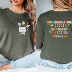 May include: Military green t-shirt with the text "COMMUNICATION LOOKS DIFFERENT FOR EVERYONE" on the back. The front features a keyboard graphic, a daisy, a speech bubble, and a hand gesture. The name "Suzy" is written below the keyboard graphic.