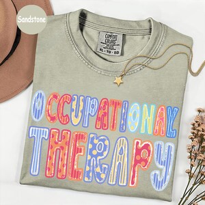 May include: A sandstone-colored short-sleeve t-shirt with a crew neckline. The shirt features the text OCCUPATIONAL THERAPY printed in large, multi-colored letters with various patterns including stripes and polka dots in blue, pink, and yellow.
