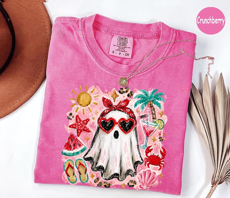 May include: A pink short-sleeve t-shirt featuring a graphic of a ghost wearing a red bandana and heart-shaped sunglasses. The design includes summer elements like a palm tree, sun, starfish, watermelon slice, flip-flops, and a cocktail.