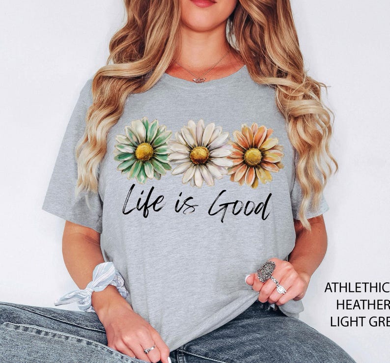 Puede incluir: Camiseta gris claro con tres flores de margarita en verde, blanco y naranja, con la frase "Life is Good" en escritura negra. La camiseta est&aacute; hecha de un material suave y jaspeado.